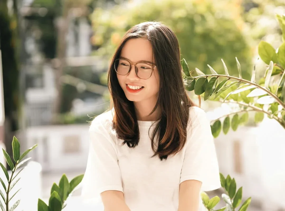 A smiling young woman in glasses and a white shirt among green leaves